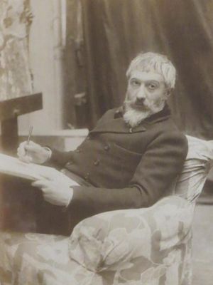 homme à la barbe blanche assis dans un fauteuil, tenant une planche à dessin et un crayon dans les main et fixant l'objectif.