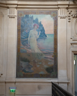 Femme en robe blanche, coiffée d'une couronne de fleurs marchant le long d'une côte rocailleuse, mer visible sur la droite de l'image.