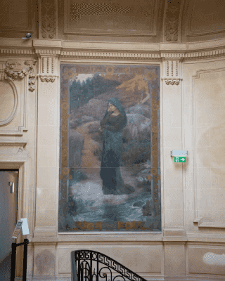 Femme habillée d'un vêtement sombre à capuche, tête basse, paysage rocheux et forestier.