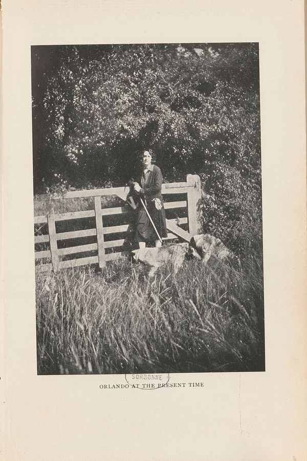 Photographie d’une femme en pied à l’orée d’une forêt. Elle se tient contre une barrière en bois qui borde un champ, deux chiens passent devant ses jambes. Elle est vêtue d’une chemise, d’un gilet et d’une jupe. Elle tient entre ses mains une canne et un chapeau. Ses cheveux sont courts.