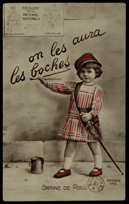 Carte photographique colorisée : un jeune enfant en robe rayée blanche et rouge et képi rouge peint un slogan au pinceau sur un mur. Il porte un petit fusil en bandoulière. Un pot de peinture est posé au sol.