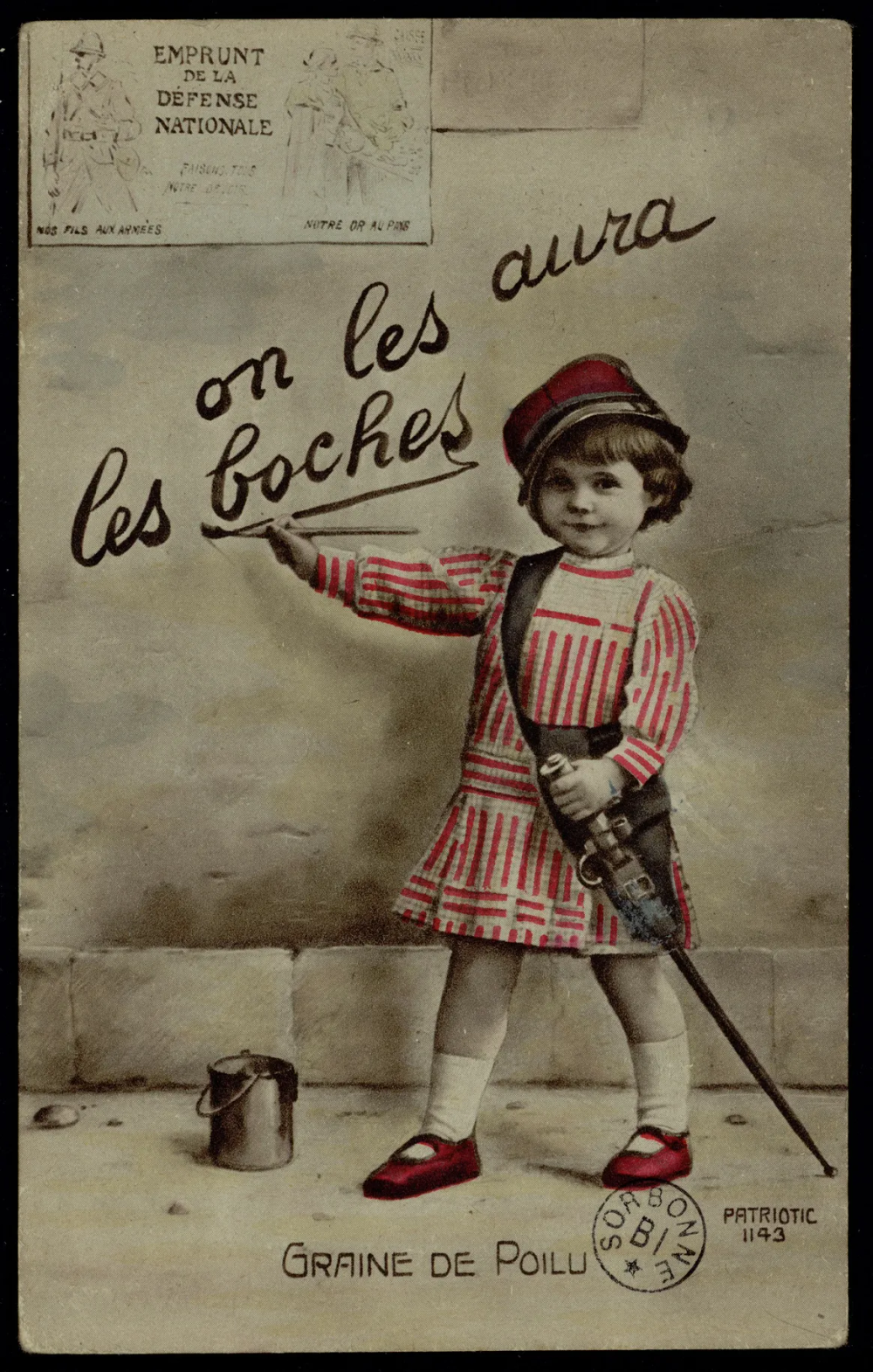 Carte photographique colorisée : un jeune enfant en robe rayée blanche et rouge et képi rouge peint un slogan au pinceau sur un mur. Il porte un petit fusil en bandoulière. Un pot de peinture est posé au sol.