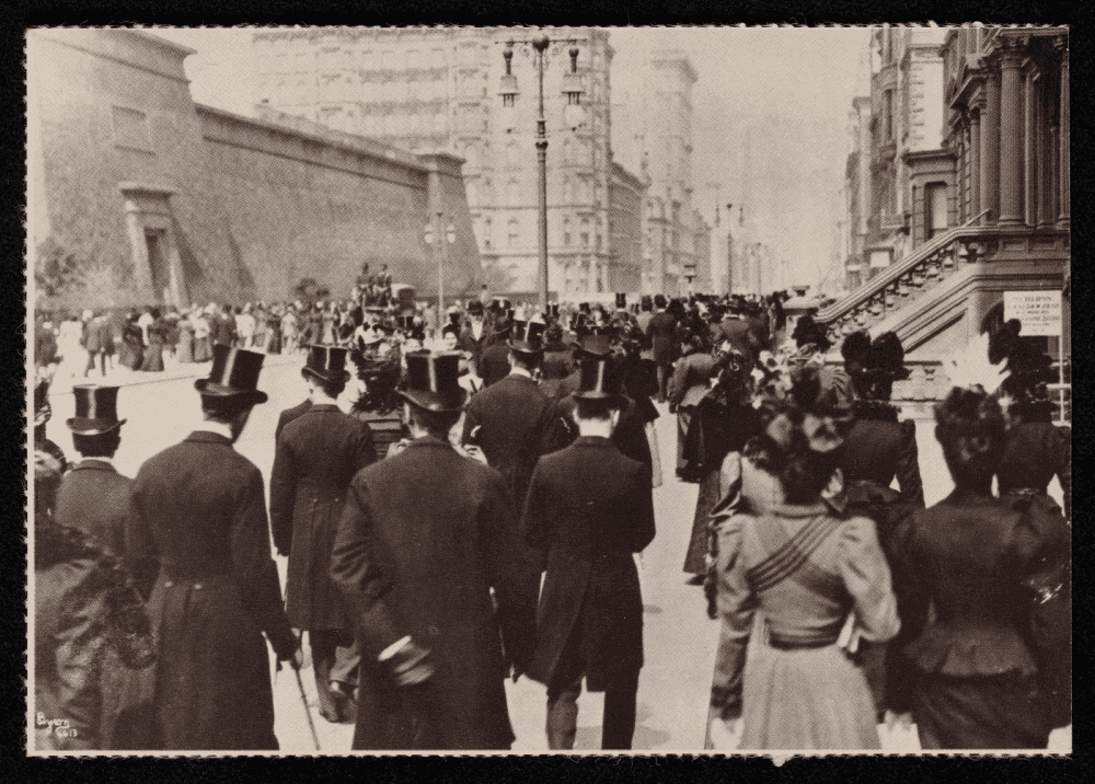 Carte postale montrant une rue urbaine du début du XXe siècle, avec hommes en hauts-de-forme et femmes en robes longues vus de dos.