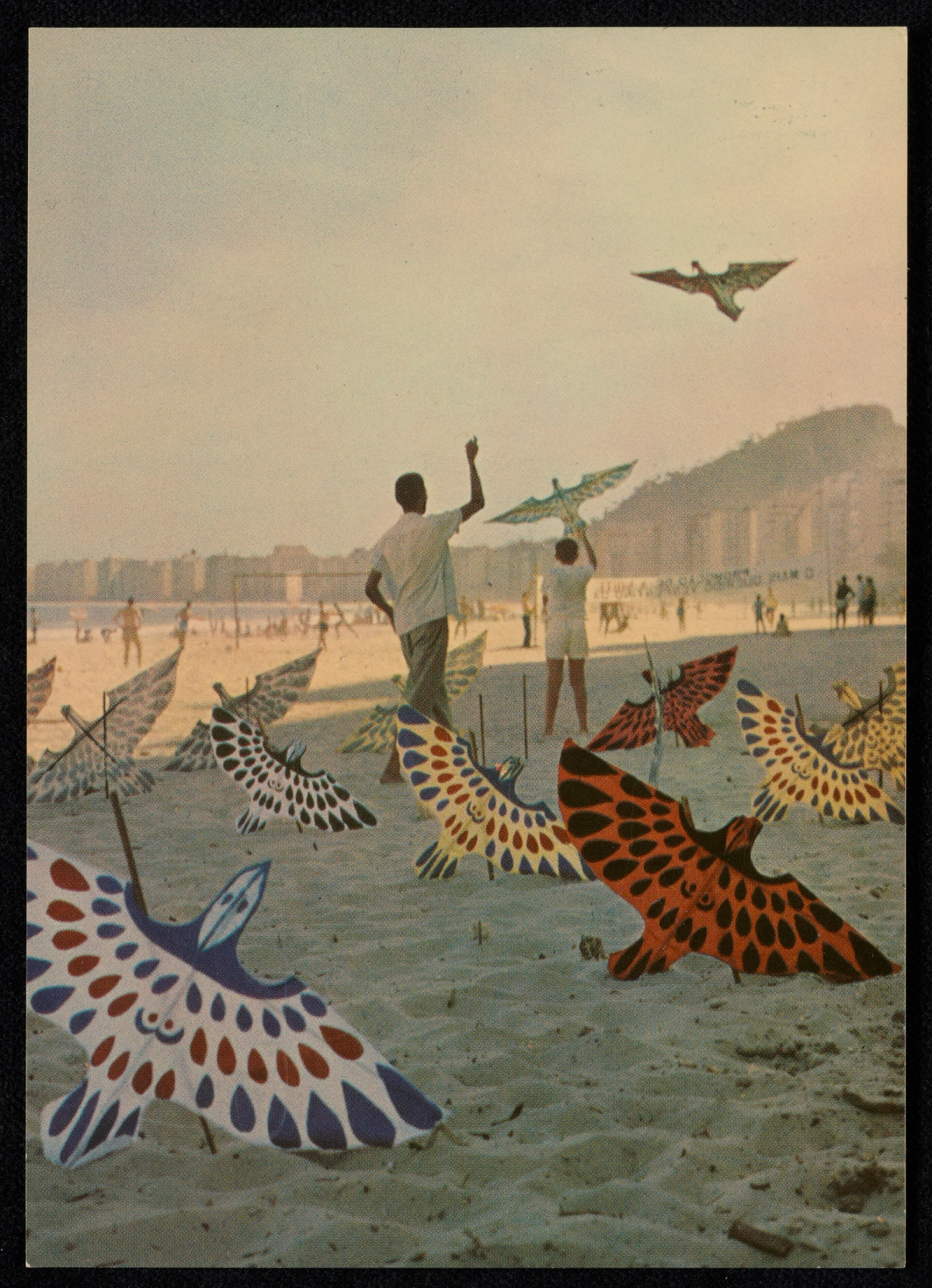 Carte postale colorée montrant une plage au sable clair où des enfants font voler des cerfs-volants en forme d'oiseaux aux ailes multicolores.