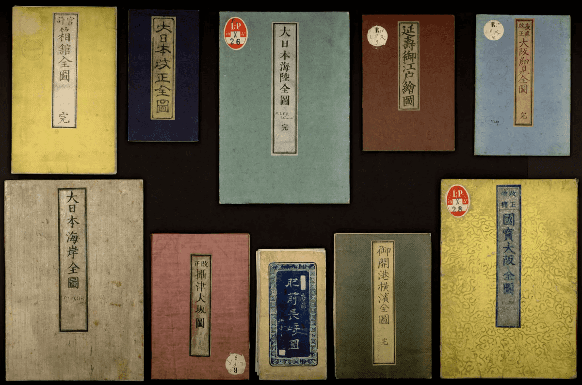 Dix couvertures cartonnées de cartes japonaises pliées, de couleurs variées, portant des titres calligraphiés en japonais et des étiquettes de cote colorées, photographiées sur fond noir.