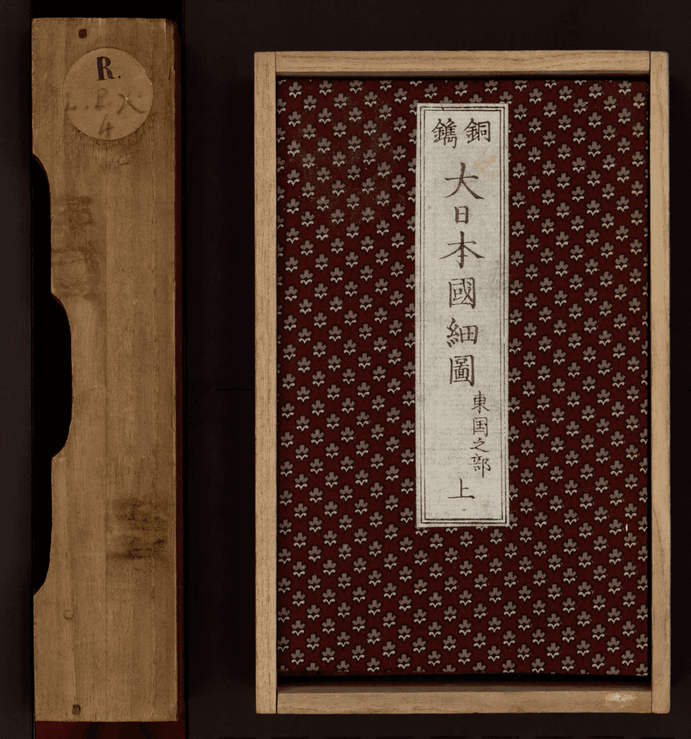 Boîte de rangement en bois clair et plat supérieur d'un atlas : couverture en tissu bordeaux à motif floral répété, pièce de titre calligraphiée à l'encre noire sur fond blanc en bandeau vertical central, étiquette de cote manuscrite sur la tranche de la boîte.