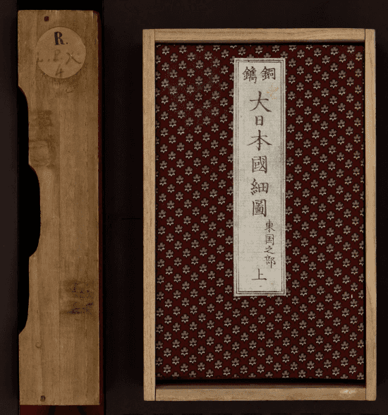 Boîte de rangement en bois clair et plat supérieur d'un atlas : couverture en tissu bordeaux à motif floral répété, pièce de titre calligraphiée à l'encre noire sur fond blanc en bandeau vertical central, étiquette de cote manuscrite sur la tranche de la boîte.