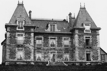 Photographie en noir et blanc : façade symétrique d’un château, deux pavillons d’angle à hauts toits, lucarnes cintrées, murs de moellons, perron central.