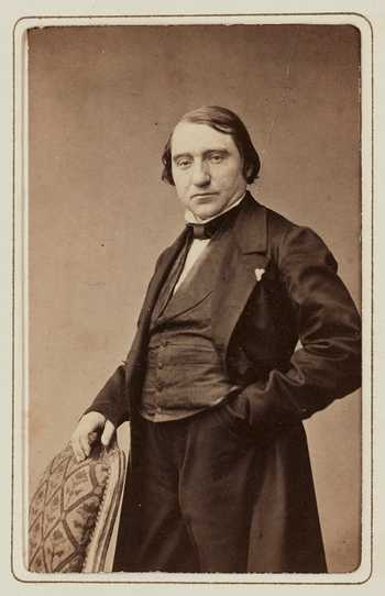 Photographie d'un homme debout vêtu à la mode du milieu du XIXe siècle avec une main posée sur le dossier d'une chaise.
