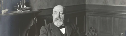 Photographie en noir et blanc d'Ernest Lichtenberger dans son bureau à la Sorbonne au début du XXe siècle. Le professeur, vêtu d'un costume sombre avec nœud papillon, est assis à son bureau sur lequel sont disposés des livres et des documents. Il arbore une barbe blanche fournie et regarde directement l'objectif. La scène est capturée dans un intérieur à boiseries typique de la Sorbonne. Au bas de l'image figure la légende : 'PARIS. - La Sorbonne, M. le Professeur E. Lichtenberger (Allemand)'. La photographie porte le numéro 1697 et est créditée 'ND Phot'.