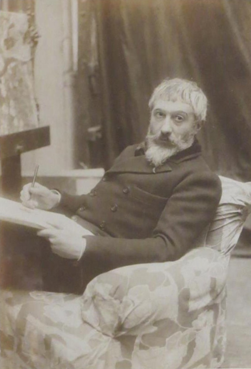 homme à la barbe blanche assis dans un fauteuil, tenant une planche à dessin et un crayon dans les main et fixant l'objectif.