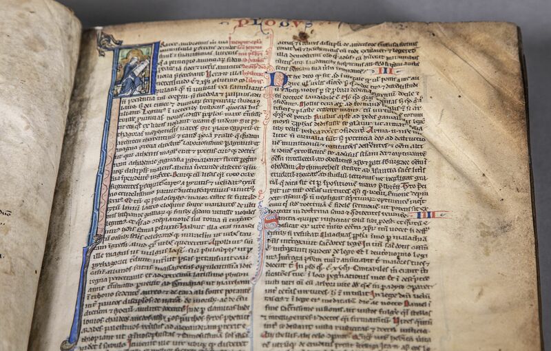 Haut du même feuillet du Ms 151 montrant de profonds plis obliques parallèles entre eux sur la partie supérieure droite, avant l'intervention de la restauratrice.  haut de la colonne gauche de texte, une lettrine P historiée montrant un homme en train d'écrire. Titre courant rouge et bleu "PROLOGUS" et lettrines filigranées en deuxième colonne.