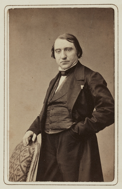 Photographie d'un homme debout vêtu à la mode du milieu du XIXe siècle avec une main posée sur le dossier d'une chaise