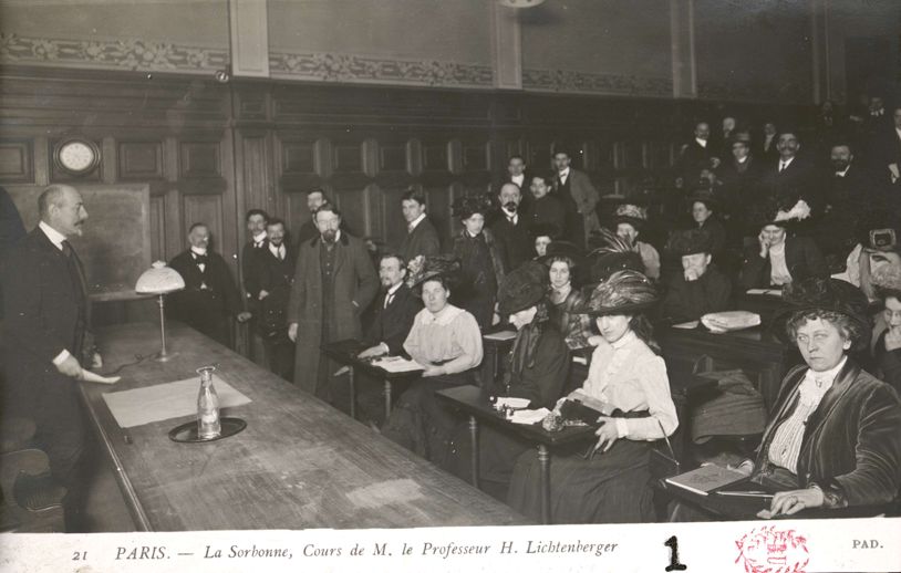 Photographie en noir et blanc d'un cours d'Henri Lichtenberger à la Sorbonne au début du XXe siècle. On y voit une salle de classe avec des boiseries ornementées typiques de la Sorbonne. Le professeur Lichtenberger se tient debout derrière son bureau sur lequel est posée une lampe. Face à lui, l'amphithéâtre est occupé par des étudiants et étudiantes, certaines portant des chapeaux élaborés caractéristiques de l'époque. Les auditeurs sont assis sur des bancs en gradins et semblent prendre des notes. La légende indique : 'PARIS. - La Sorbonne, Cours de M. le Professeur H. Lichtenberger'. La photographie est créditée 'PAD.' et porte le numéro 21.