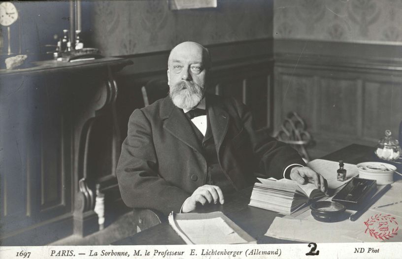 Photographie en noir et blanc d'Ernest Lichtenberger dans son bureau à la Sorbonne au début du XXe siècle. Le professeur, vêtu d'un costume sombre avec nœud papillon, est assis à son bureau sur lequel sont disposés des livres et des documents. Il arbore une barbe blanche fournie et regarde directement l'objectif. La scène est capturée dans un intérieur à boiseries typique de la Sorbonne. Au bas de l'image figure la légende : 'PARIS. - La Sorbonne, M. le Professeur E. Lichtenberger (Allemand)'. La photographie porte le numéro 1697 et est créditée 'ND Phot'.